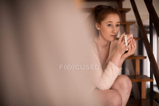 Задумчивая женщина пьет кофе, расслабляясь дома на лестнице — стоковое фото