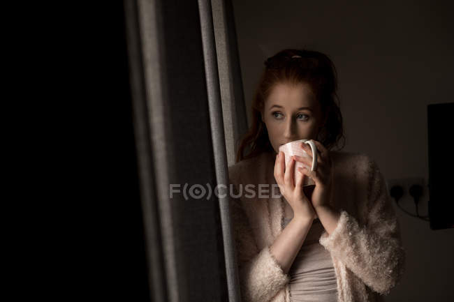 Задумчивая женщина пьет кофе, глядя в окно дома — стоковое фото