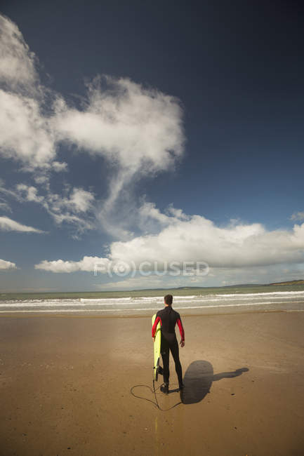Surfista con tabla de surf mirando al mar desde la playa en un día soleado - foto de stock