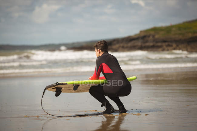 Surfista com prancha de surf agachado na praia em um dia ensolarado — Fotografia de Stock