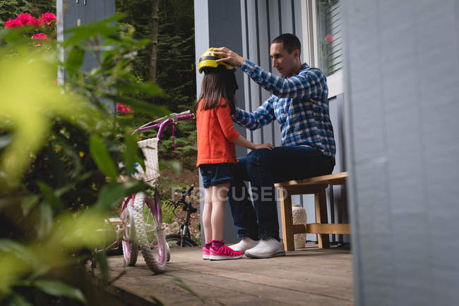Vater hilft seiner Tochter, Fahrradhelm auf der Veranda aufzusetzen — Stockfoto