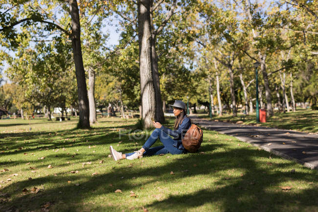 Mujer joven sentada en el parque usando teléfono móvil - foto de stock
