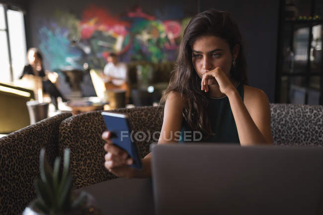 Красивая деловая женщина с помощью смартфона во время работы на ноутбуке в офисной столовой — стоковое фото