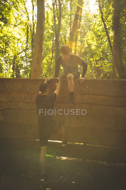 Un homme apte à aider son coéquipier à escalader un mur en bois au camp d'entraînement — Photo de stock