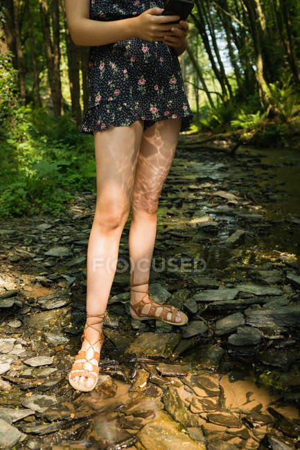 Low section of woman standing near the river coast and using mobile phone in green forest — Stock Photo