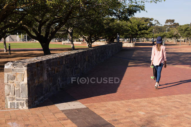 Вид ззаду жінки, що йде в парку зі скейтбордом — стокове фото