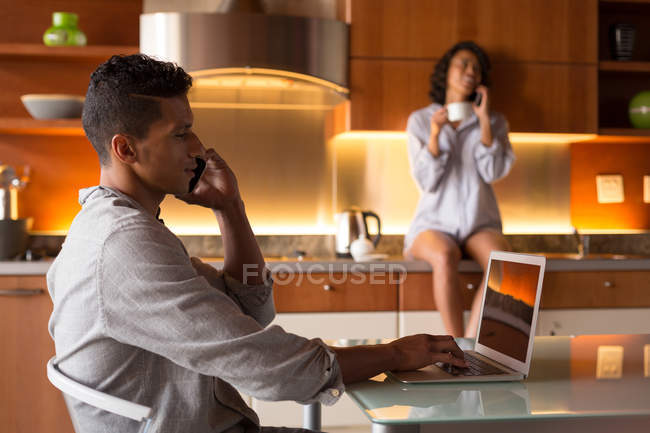 Homme parlant sur un téléphone mobile tout en utilisant un ordinateur portable dans la cuisine à la maison — Photo de stock