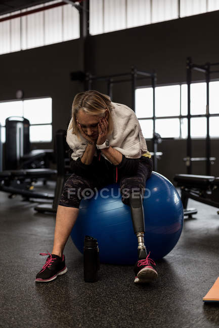 Mujer discapacitada preocupada sentada en una pelota de ejercicio en el gimnasio - foto de stock