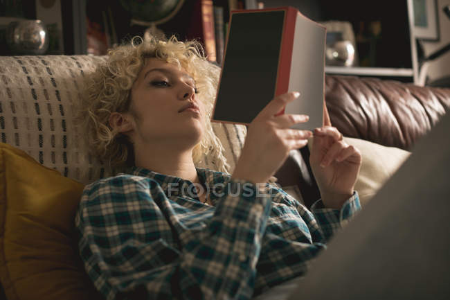 Jovem mulher lendo um livro na sala de estar em casa — Fotografia de Stock