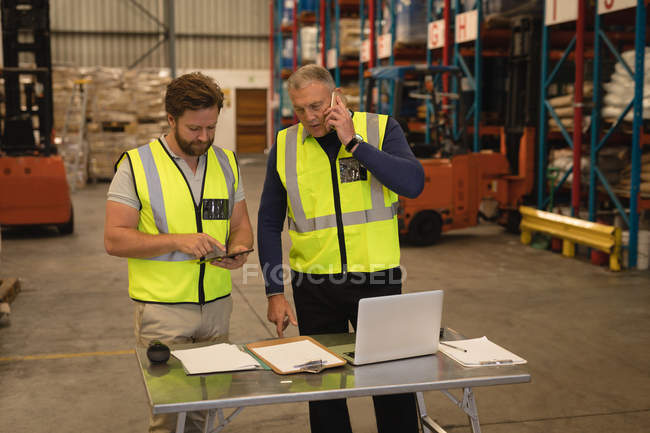 Supervisores masculinos hablando por teléfono móvil y utilizando tableta digital en el almacén - foto de stock