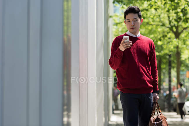 Jeune homme utilisant un téléphone portable tout en marchant sur le trottoir — Photo de stock