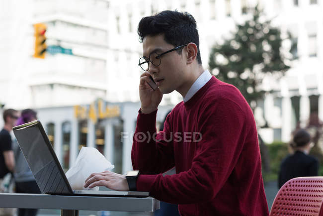 Hombre joven hablando en el teléfono móvil mientras usa el ordenador portátil en la cafetería al aire libre - foto de stock