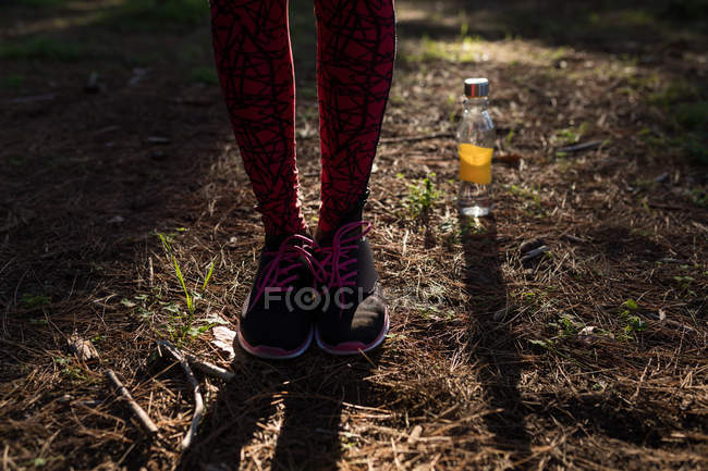 Sección baja de la atleta femenina con zapatillas de deporte de pie junto a la botella de agua en el bosque - foto de stock