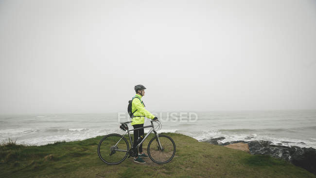 Young man with cycle standing on hill at sea coast — Stock Photo
