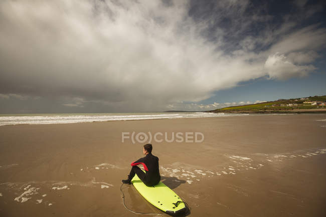 Surfeur assis sur la planche de surf sur la plage et regardant la mer par une journée ensoleillée — Photo de stock