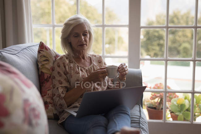 Mujer mayor tomando café mientras usa el portátil en el sofá en casa - foto de stock
