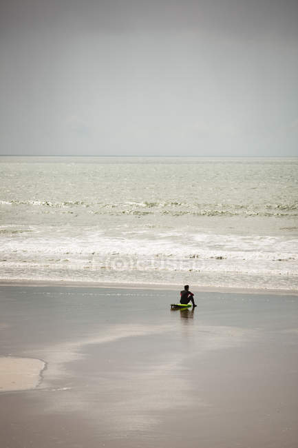 Surfista sentado en la tabla de surf en la playa y mirando al mar en un día soleado - foto de stock