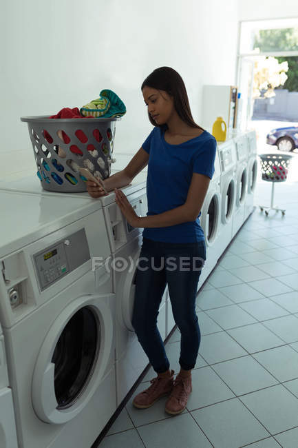 Jeune femme utilisant son téléphone portable à la laverie automatique — Photo de stock