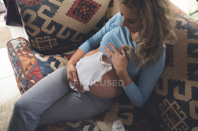 Mulher grávida com roupas de bebê na sala de estar em casa — Fotografia de Stock