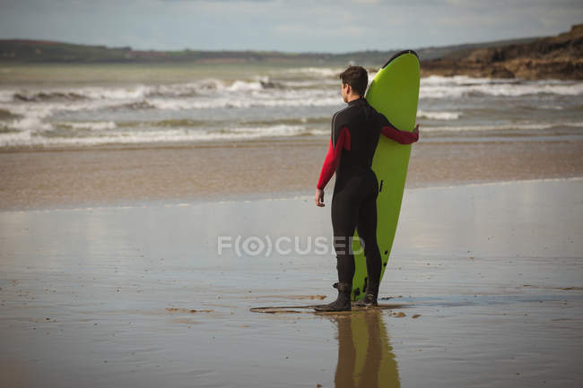 Surfista con tabla de surf mirando al mar desde la playa - foto de stock