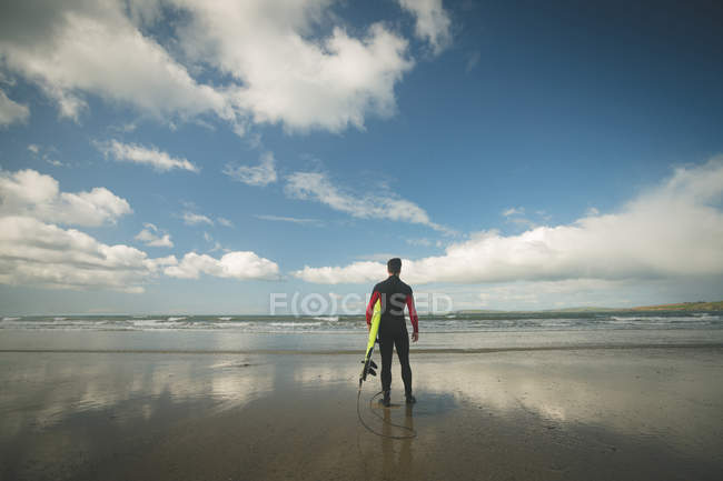 Вид сзади серфингиста с доской для серфинга, смотрящего на море с пляжа — стоковое фото