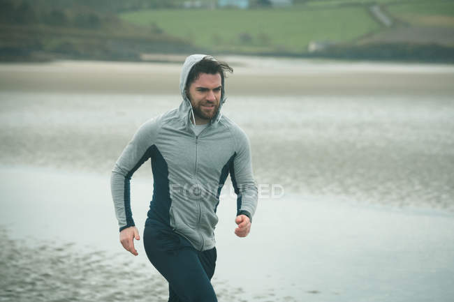 Fit man man in hoodie jogging on beach — Stock Photo