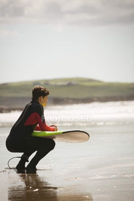 Surfista com prancha de surf agachado na praia em um dia ensolarado — Fotografia de Stock