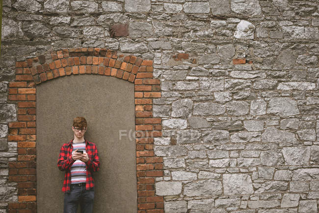 Joven parado contra la pared de piedra y usando su teléfono móvil - foto de stock