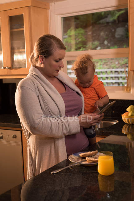 Mutter mit Baby steht in der Küche und nutzt digitales Tablet zu Hause — Stockfoto