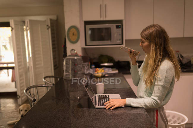 Chica tomando en el teléfono móvil mientras se utiliza el ordenador portátil en casa - foto de stock