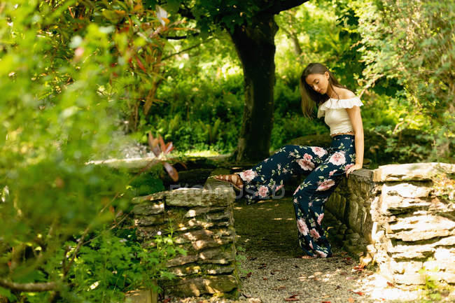 Hermosa mujer sentada en una pared de piedra en el bosque verde en un día soleado - foto de stock