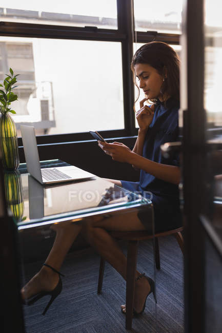 Бізнес-леді сидить і використовує телефон під час роботи на ноутбуці в офісі — стокове фото
