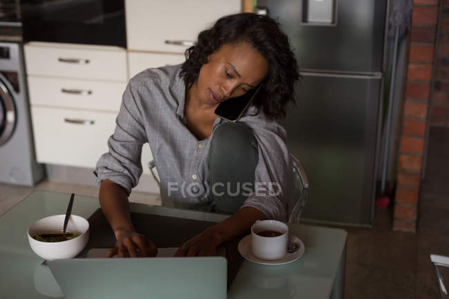 Donna che parla sul telefono cellulare mentre utilizza il computer portatile a casa — Foto stock