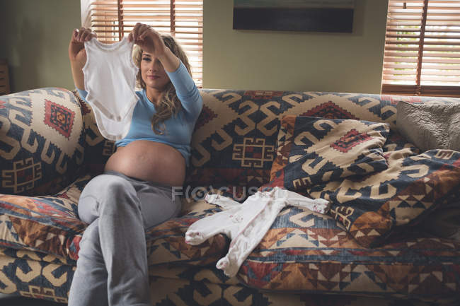 Mujer embarazada mirando ropa de bebé en la sala de estar en casa - foto de stock