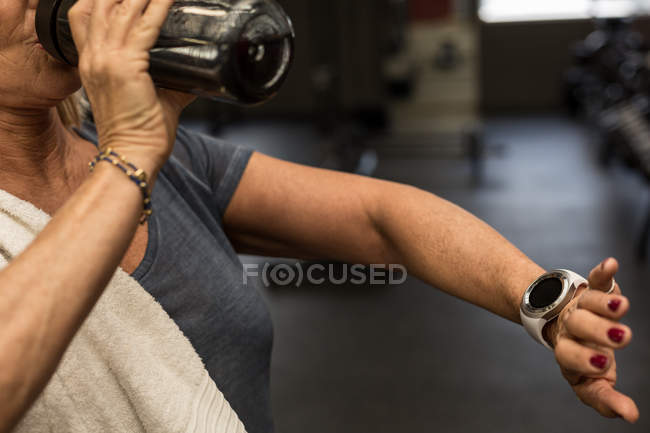 Seção média de mulher madura verificando o tempo enquanto bebe água no ginásio — Fotografia de Stock