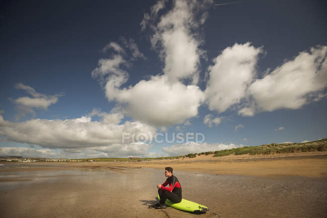 Surfer sentado en la tabla de surf y mirando al mar en la playa - foto de stock