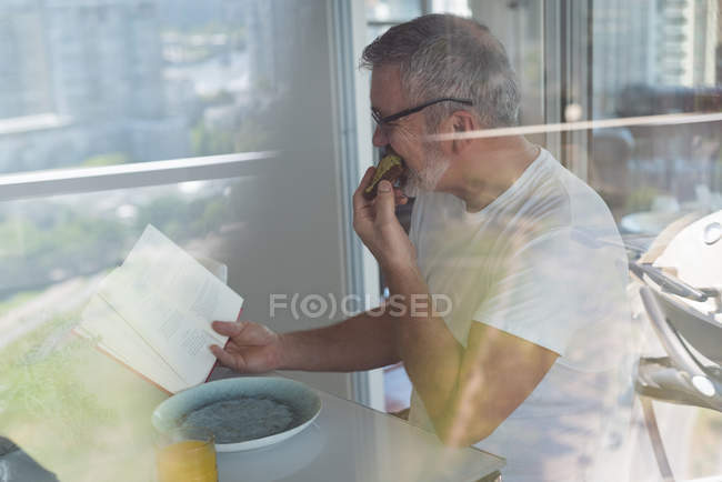 Homem lendo um livro enquanto toma café da manhã em casa — Fotografia de Stock