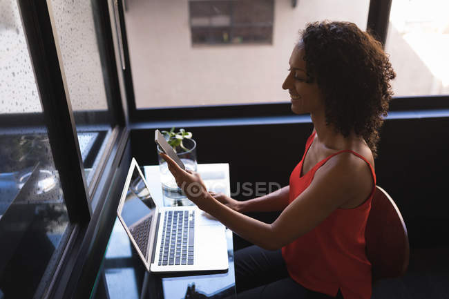 Femme d'affaires assis et utilisant le téléphone tout en travaillant sur ordinateur portable au bureau — Photo de stock