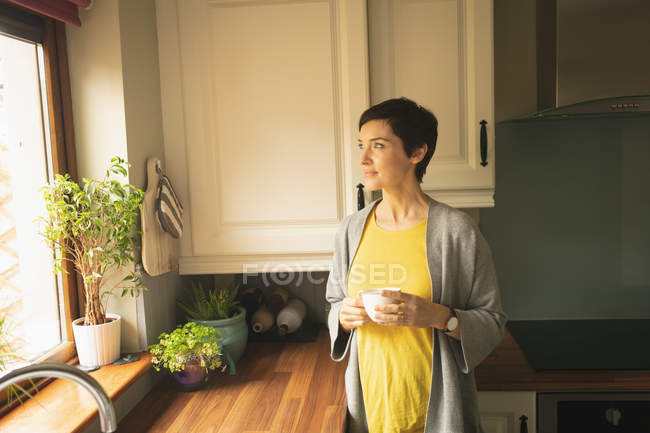 Mulher tomando café olhando através da janela na cozinha em casa — Fotografia de Stock