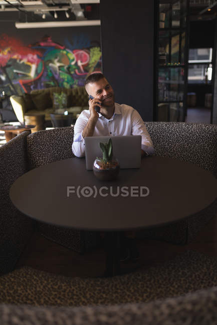 Empresário falando por telefone enquanto trabalhava em laptop no refeitório do escritório — Fotografia de Stock