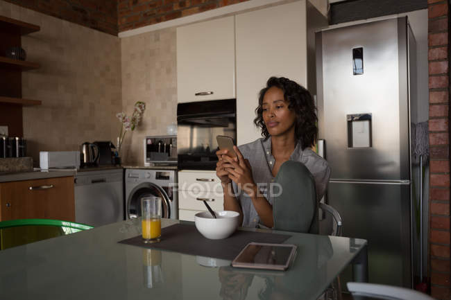 Mujer usando el teléfono móvil mientras desayuna en casa - foto de stock