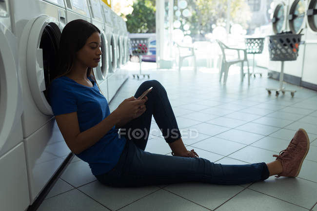 Jovem mulher sentada no chão e usando seu telefone na lavanderia — Fotografia de Stock
