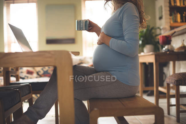 Femme enceinte utilisant un ordinateur portable tout en prenant un café à la maison — Photo de stock