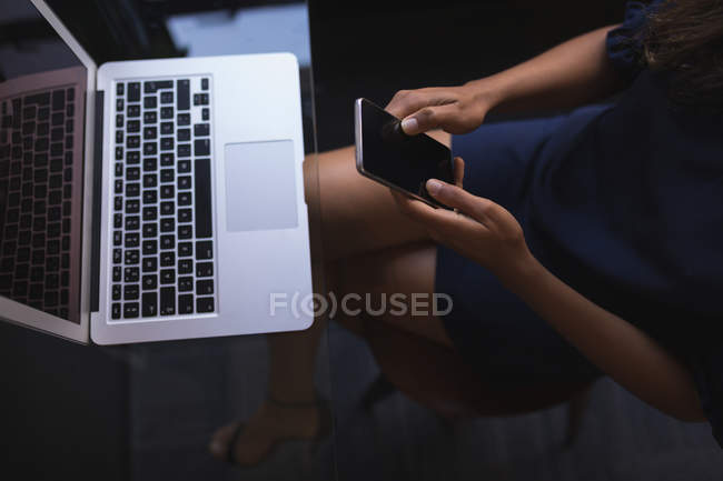 Empresária sentada e usando telefone enquanto trabalhava no laptop no escritório — Fotografia de Stock