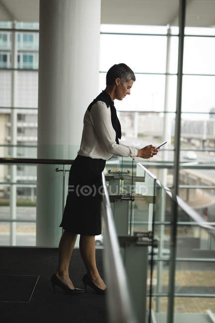 Бизнесмен, использующая свой мобильный телефон возле перила в офисе — стоковое фото
