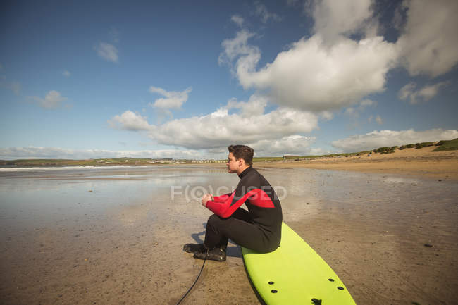 Vista lateral do surfista sentado na prancha de surf na praia e olhando para o mar — Fotografia de Stock