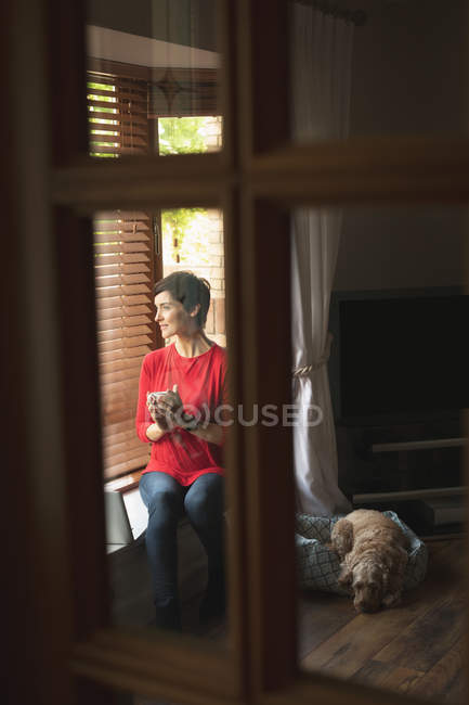 Donna che guarda attraverso la finestra mentre prende un caffè in soggiorno a casa — Foto stock