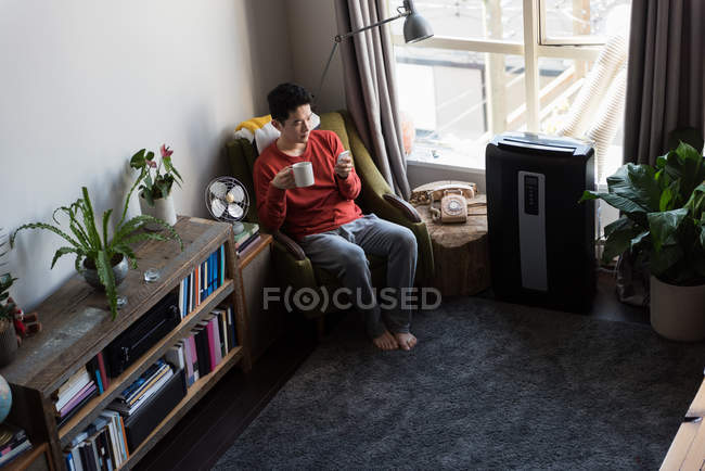 Uomo che prende il caffè mentre utilizza il telefono cellulare in soggiorno a casa — Foto stock