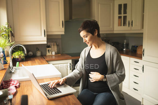 Pregnant woman using laptop in kitchen at home — Stock Photo