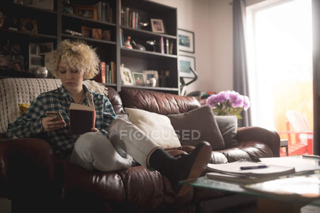 Femme utilisant un téléphone portable tout en lisant le livre dans le salon à la maison — Photo de stock